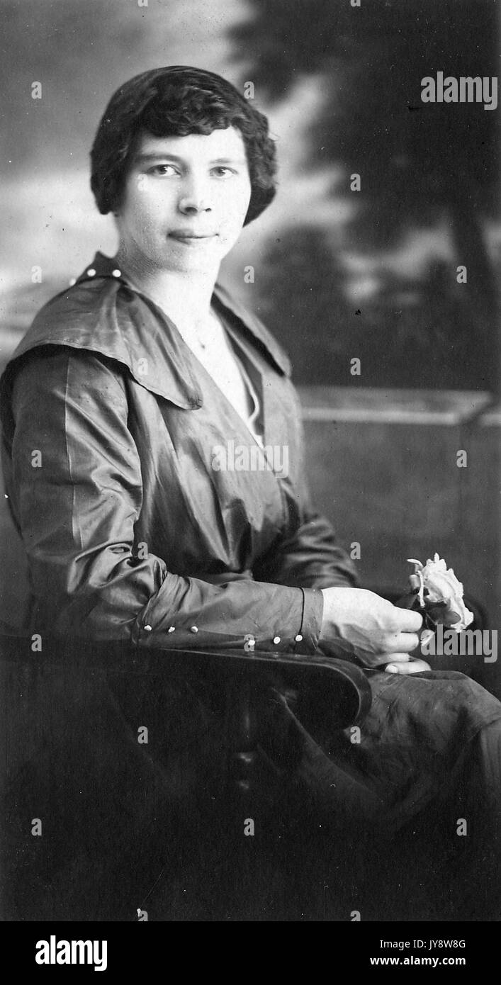 Drei viertel länge Portrait von Sitzen afrikanische amerikanische Frau, mit ernsten Gesichtsausdruck, trug ein dunkles Kleid, in einem hölzernen Stuhl sitzen, mit einer Blume in der Hand, 1915. Stockfoto