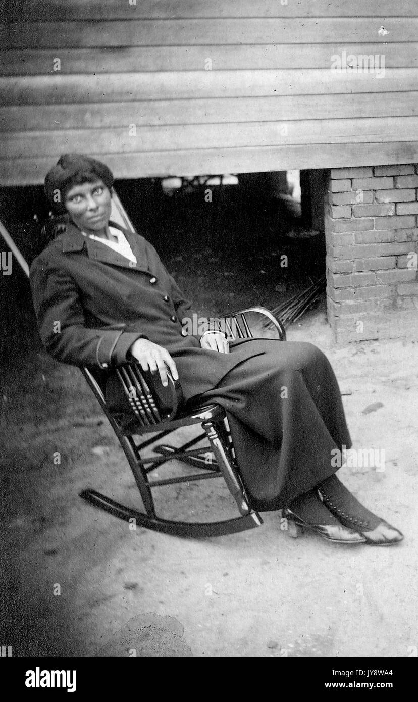 In voller Länge Porträt der Afrikanische amerikanische Frau, die in eine Holzkiste Schaukelstuhl sitzt, lehnte sich zurück, mit einem ernsten Gesichtsausdruck, trug ein dunkles Kleid, 1920. Stockfoto