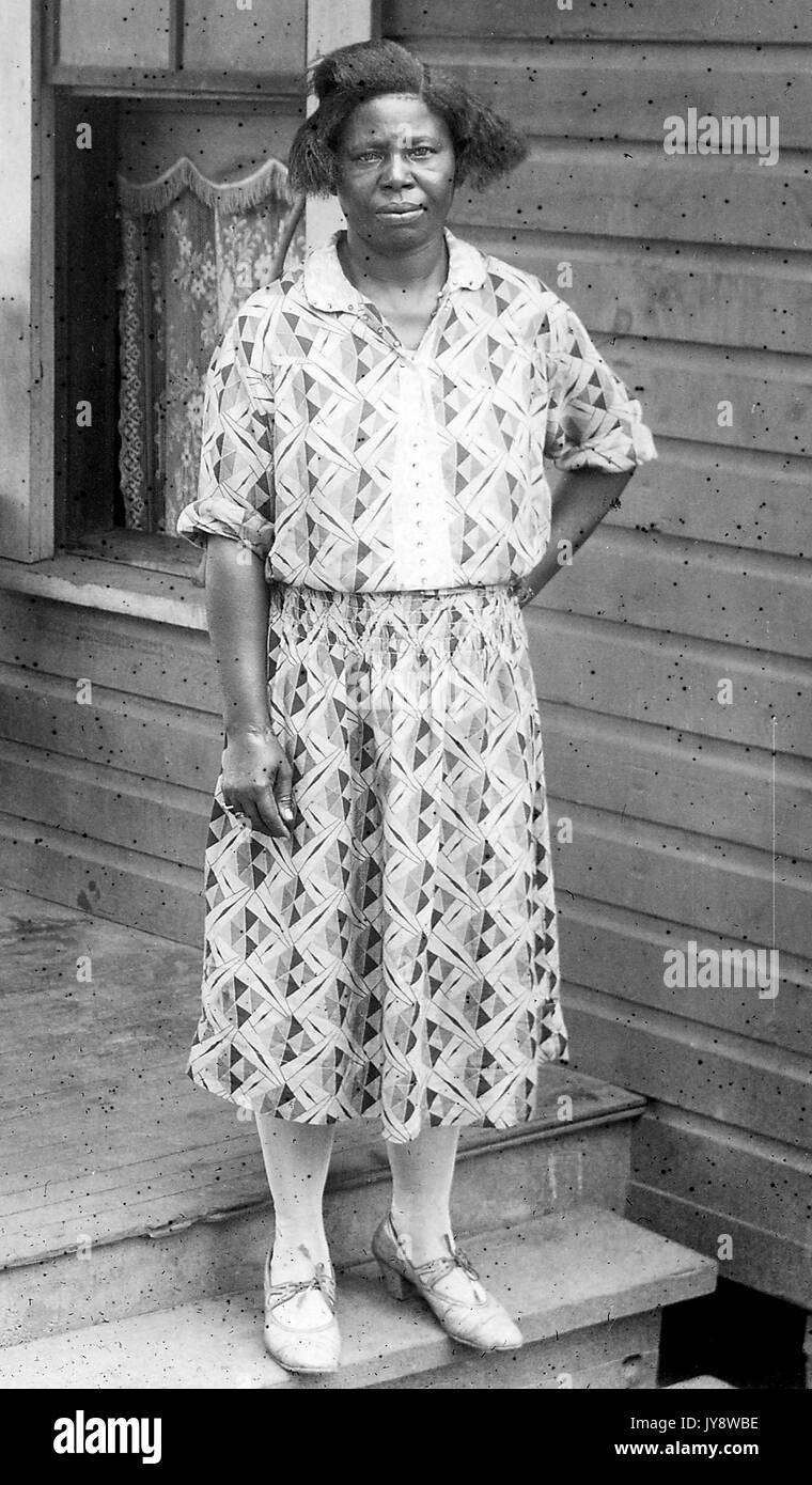In voller Länge Porträt der Afrikanische amerikanische Frau, stehend auf einer Veranda, eine Hand auf ihre Hüfte, trägt ein gemustertes Kleid, mit einem ernsten Gesichtsausdruck, 1930. Stockfoto