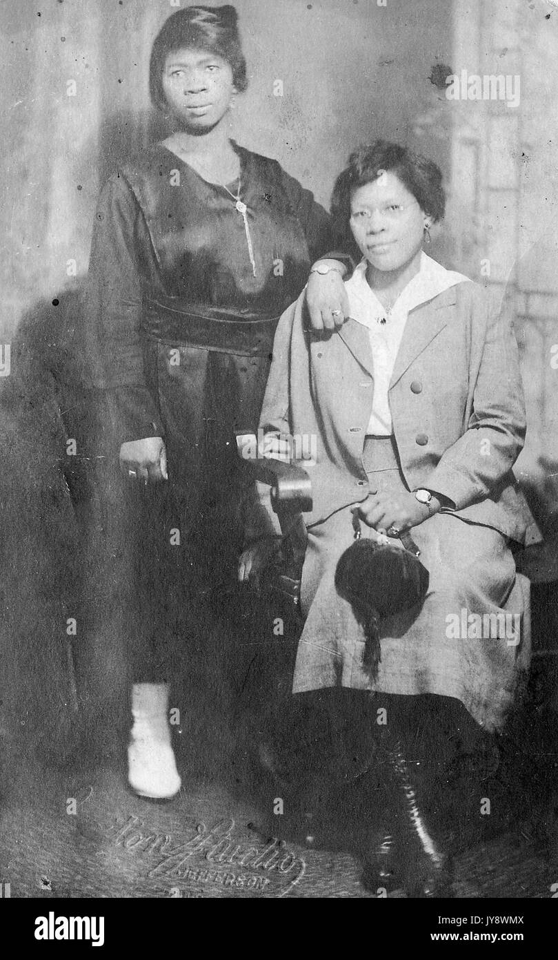 In voller Länge Porträt von zwei afrikanische amerikanische Frauen, einer ständigen mit ihrem Arm auf der anderen ruhte, trägt einen dunklen Kleid mit einer langen Kette, während die andere sitzt mit einer kleinen Tasche in der einen Hand, 1920. Stockfoto