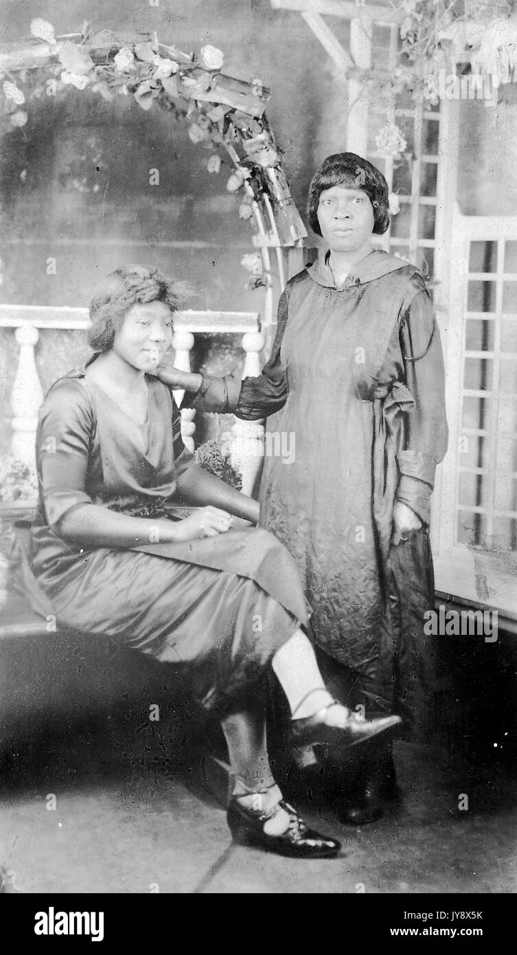 In voller Länge Porträt von zwei Afrikanische amerikanische Frauen, die Frau auf der linken Seite sitzen, mit einem grinsenden Gesichtsausdruck und gekreuzten Beinen, die andere Frau, die ihr mit der einen Hand auf die Schulter der sitzende Frau, mit einem überraschten Gesichtsausdruck, beide Frauen tragen dunkle Kleider, 1920. Stockfoto