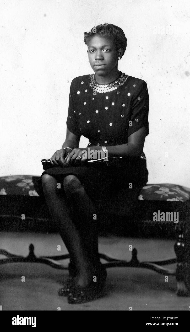 Volle Länge sitzt Porträt der jungen Afrikanische amerikanische Frau im dunklen, aufwendige Kleidung, das Tragen von Uhren und Schmuck, Geldbeutel, neutralen Ausdruck, 1920. Stockfoto