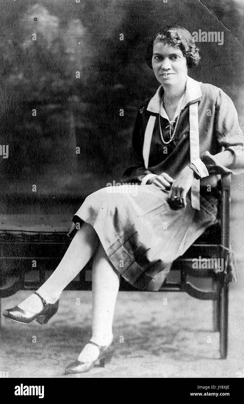Volle Länge sitzen Porträt, junge afrikanische amerikanische Frau, dunklen Kleid mit Jacke, Halskette und Schal, neutralen Gesichtsausdruck, 1920. Stockfoto