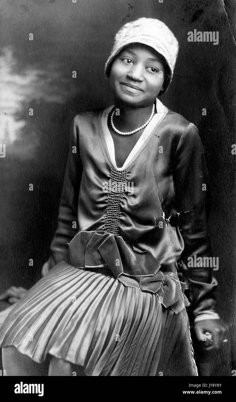 Drei Viertel Länge Porträt von afroamerikanischen Frau tragen langes Kleid, Perlenkette und Hut, lächelnd Gesichtsausdruck, Kopf geneigt, Blick auf die Seite, 1920. Stockfoto