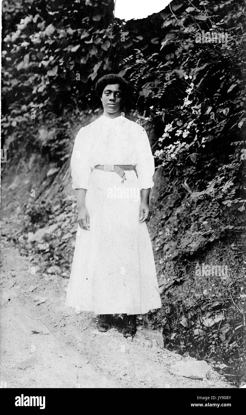 Ganzlanges Standbild einer jungen afroamerikanischen Frau, in hellem Kleid mit dunklem Gürtel, vor einer mit Pflanzen und Reben bedeckten Wand stehend, neutraler Ausdruck, 1910. Stockfoto