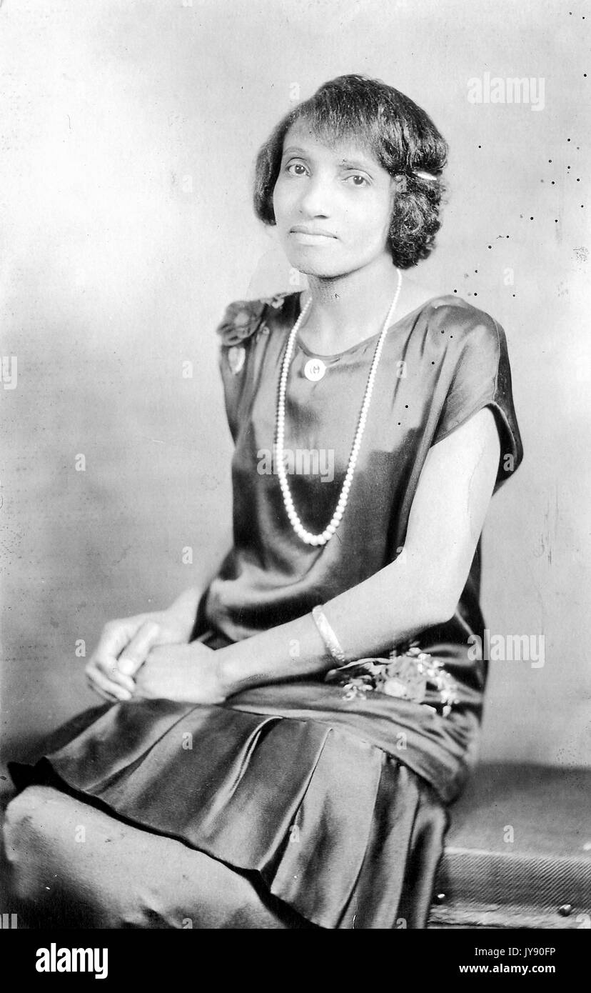 Portrait einer afrikanischen amerikanischen Frau sitzt mit ihr die Hände in ihrem Schoß gefaltet, trug ein Kleid und eine Perle Halskette, 1915. Stockfoto