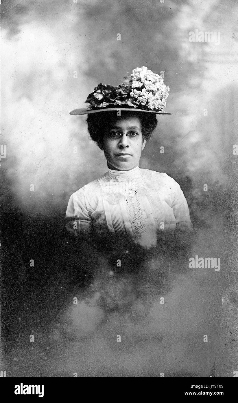 Afro-amerikanische Frau, Taille, Porträt mit einem ernsten Gesichtsausdruck, das Tragen von reich verzierten Hut mit Blumen, weißen Kleid und Gläser, 1915. Stockfoto