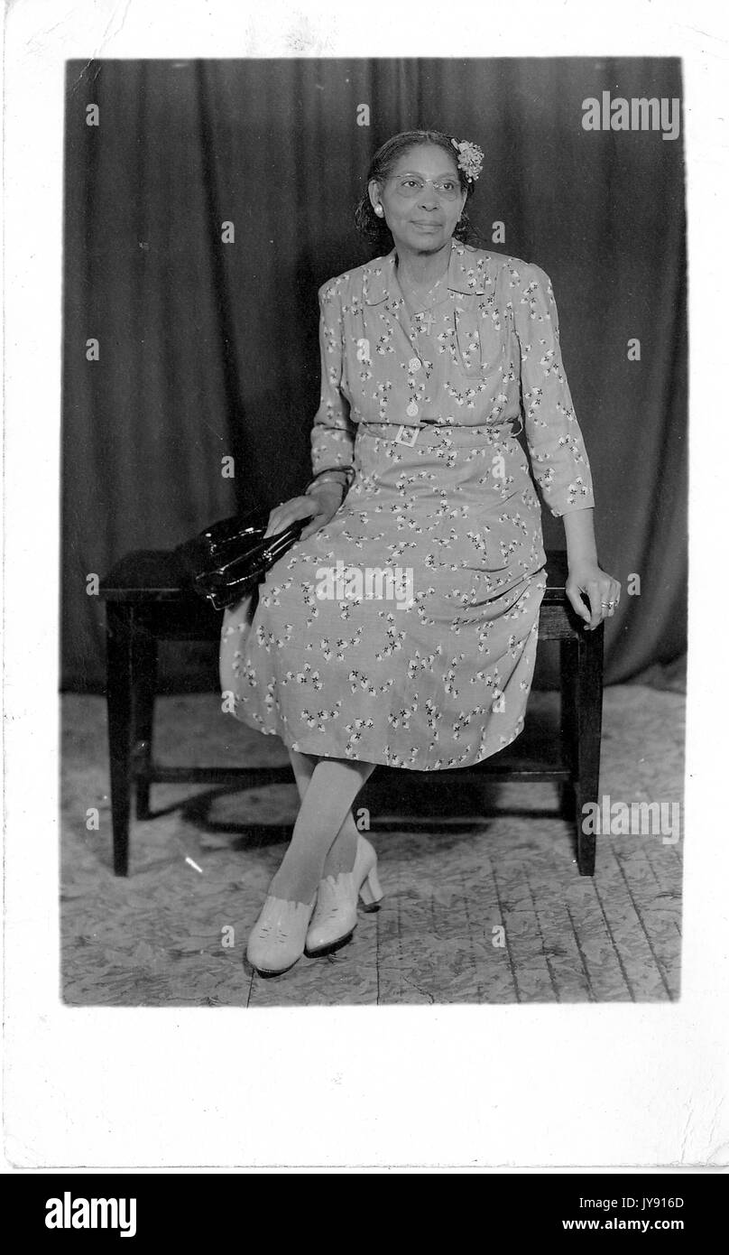 In voller Länge Porträt der afroamerikanischen Frau sitzt auf einem Holztisch, trägt ein gemustertes Kleid und weiße Schuhe und eine schwarze Handtasche, 1920. Stockfoto