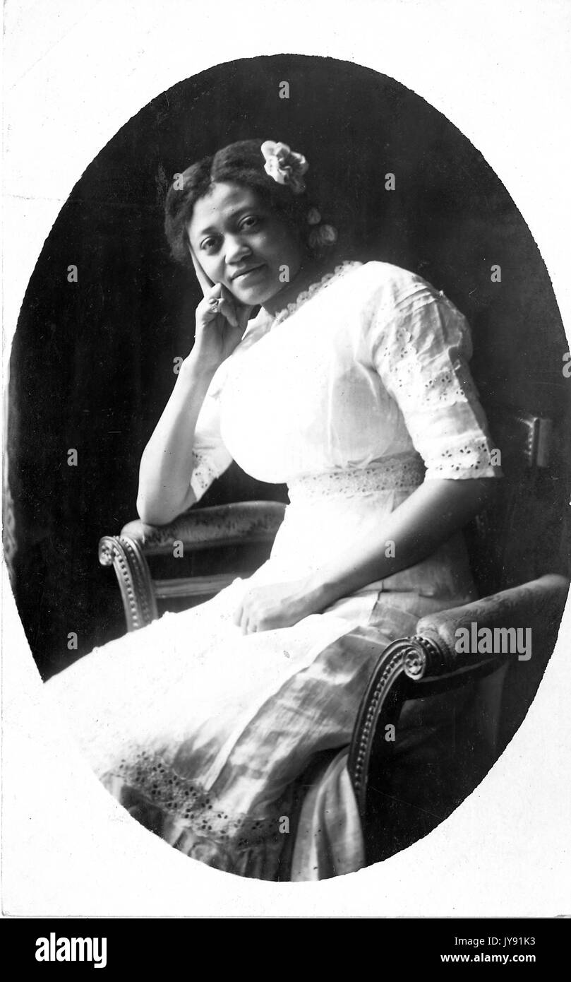 Dreiviertel sitzendes Porträt einer lächelnden jungen afroamerikanischen Frau, die ein leichtes Kleid und eine Blume im Haar trägt, den Kopf auf die rechte Hand gekippt, im gemusterten Stuhl sitzend, 1915. Stockfoto
