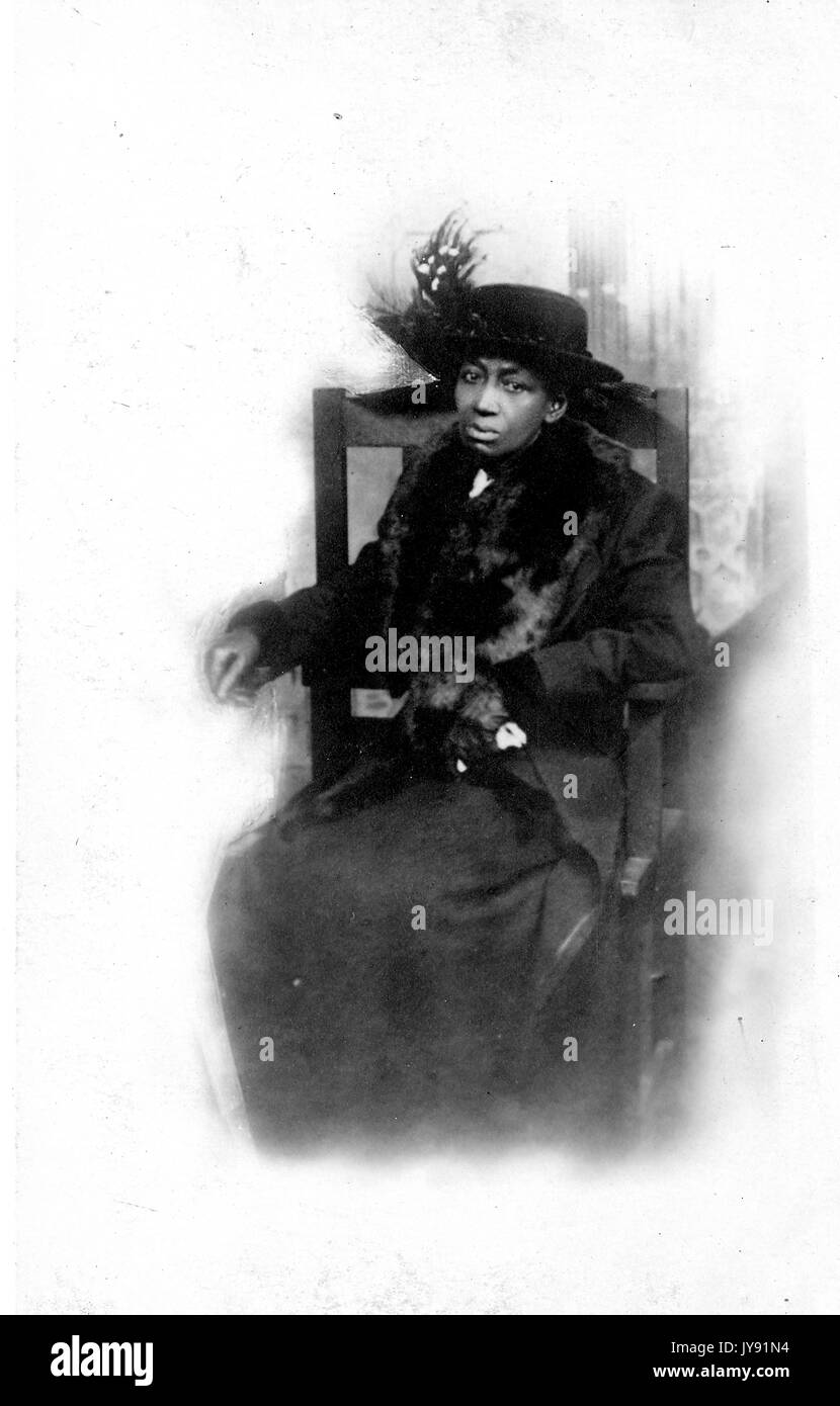 Volle Länge sitzt Portrait von Reifen afrikanische amerikanische Frau mit einem neutralen Ausdruck, trug ein dunkles Kleid, einen dunklen Mantel und Hut mit Federn, 1915. Stockfoto