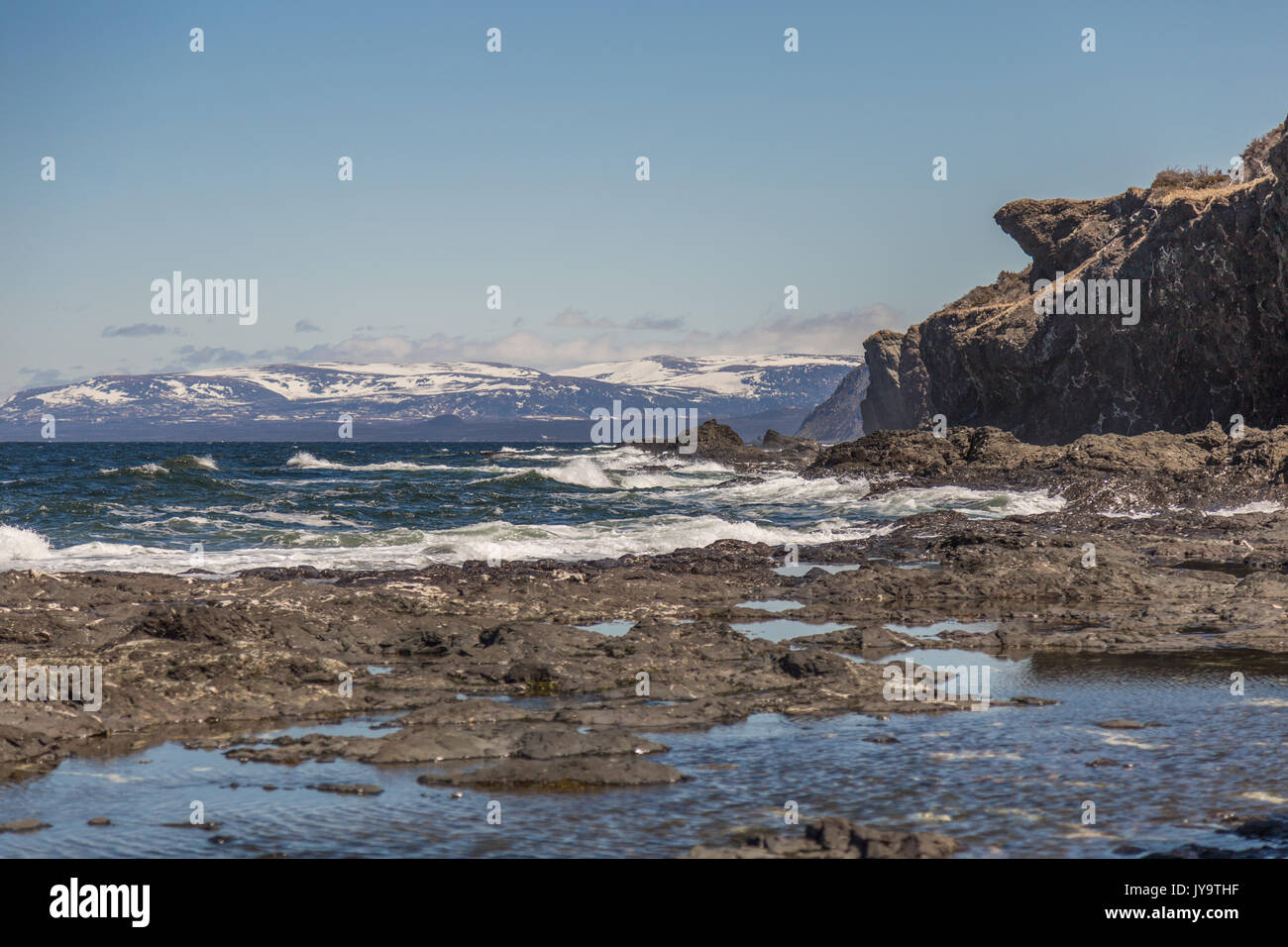 Die schneebedeckten Berge in der Ferne im Sonnenuntergang vor einem klaren blauen Himmel. White rapids und Felsen im Meer. Große schroffen Klippen. Gros Morne National P Stockfoto