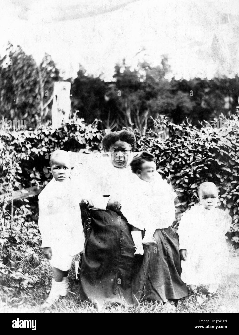 Die afroamerikanische Großmutter sitzt und hält eines von drei Enkelkindern, die anderen beiden Enkelkinder sind an ihrer Seite, sie sitzen und stehen vor Gebüsch, die Kinder tragen helle Outfits und die Großmutter trägt ein helles Hemd und einen dunklen langen Rock, 1920. Stockfoto