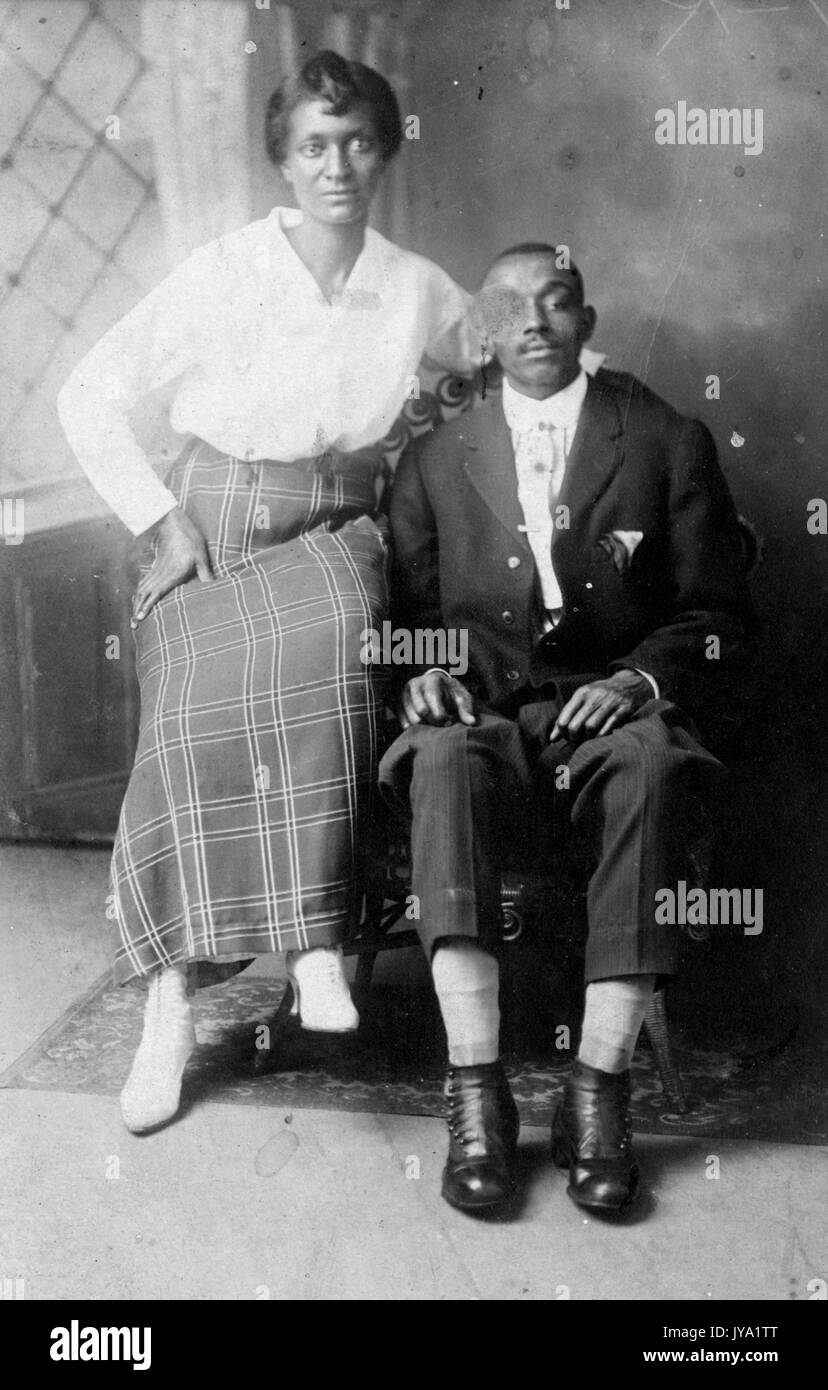 Porträt eines afroamerikanischen Mannes und seiner Frau, der Mann sitzt auf einem Stuhl vor einem dekorativen Wandgemälde und die Frau sitzt auf einem Arm des Stuhls, Der Ehemann trägt einen dunklen Anzug mit einem hellen Hemd darunter und die Frau trägt einen bodenlangen gemusterten Rock und ein hellfarbiges Hemd, 1920. Stockfoto