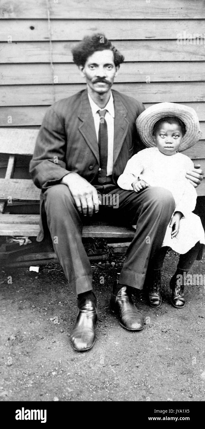 Porträt eines älteren afroamerikanischen Mannes, der mit einem kleinen Kind auf einer Bank sitzt, einen dunklen Anzug trägt und einen Arm um das junge Mädchen, das ein helles Kleid und einen Hut trägt, 1920. Stockfoto