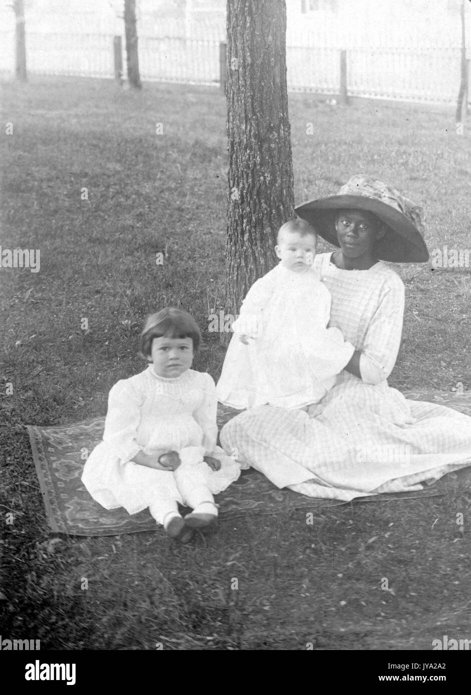 Eine afroamerikanische Kindermädchen trägt ein helles Kleid und einen großen Hut, sie sitzt auf einem Teppich auf dem Gras hält ein kaukasisches Baby und sitzt neben einem kaukasischen jungen Mädchen, sie sind alle sitzen vor einem Baum, 1920. Stockfoto