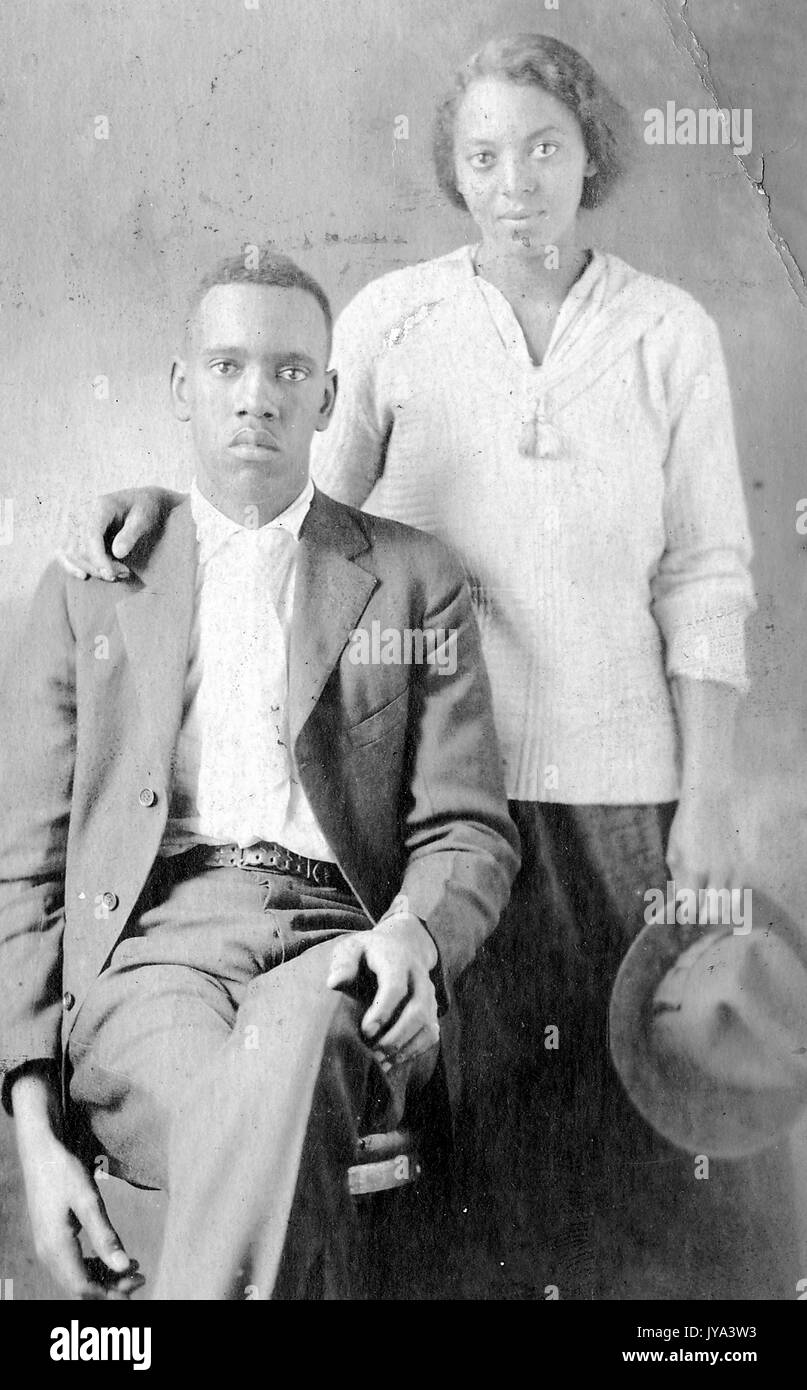 Junge afrikanische - amerikanisches Paar, der Mann sitzt auf einem Stuhl in Anzug, mit einem leeren Gesichtsausdruck, die Frau hinter ihn mit ihrer Hand auf die Schulter und halten Sie einen Hut, mit einem leichten Lächeln, 1915. Stockfoto