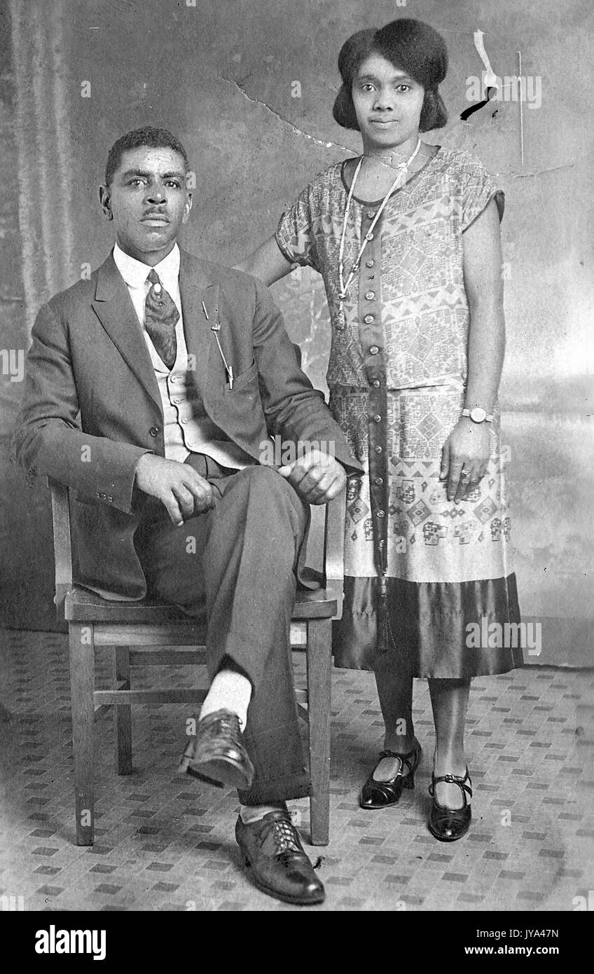 Junge afrikanische amerikanische Paar, ein Mann, der auf einem Stuhl sitzt und in Anzug, stehende Frau und trug ein Kleid, Beobachten und Lackschuhe mit ihrer Hand ruht auf dem Stuhl, auf dem Fliesenboden mit einem Hintergrund in einem Fotostudio, Ohio, 1932. Stockfoto
