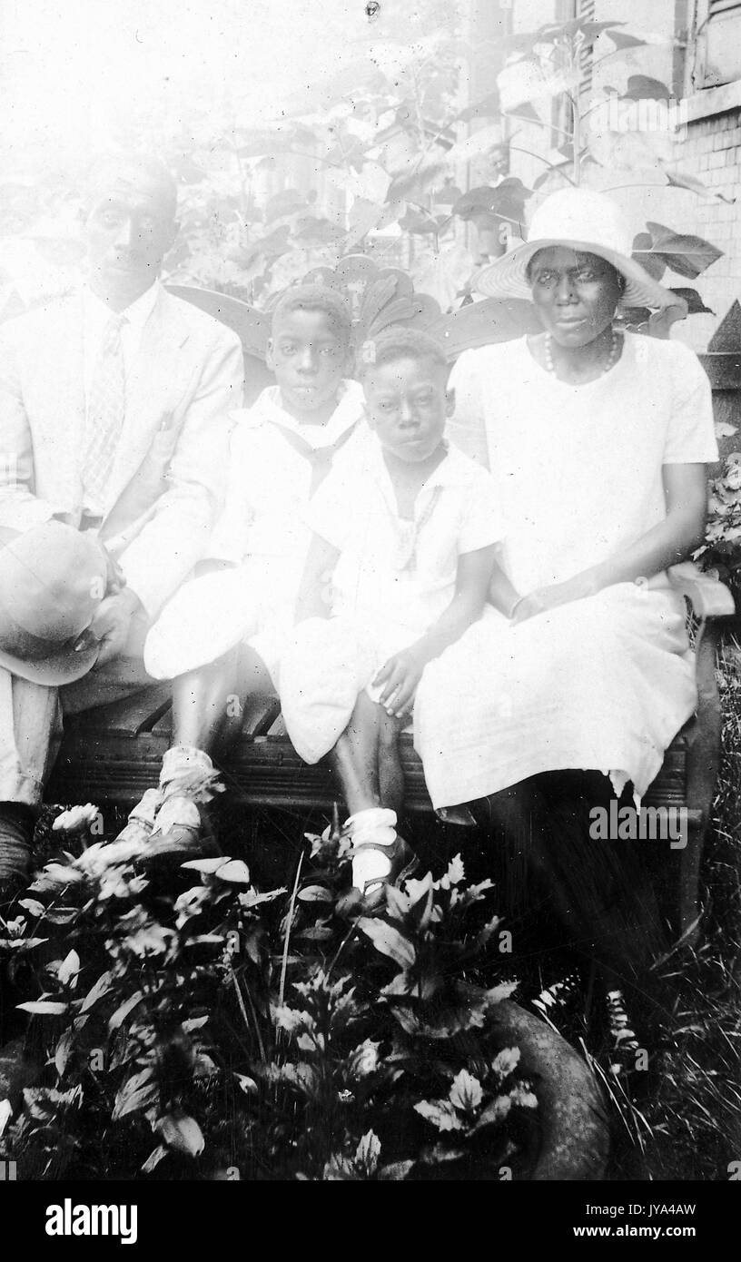 Familie der afrikanisch-amerikanischen Landarbeiter, auf einer Bank in einer Umgebung mit Pflanzen im Hintergrund sitzt ein Mann und eine Frau mit zwei Jungs, der Mann mit dem Hut in der Hand und die Frau in weißem Kleid, original Foto sehr überbelichtet ist, 1932. Stockfoto