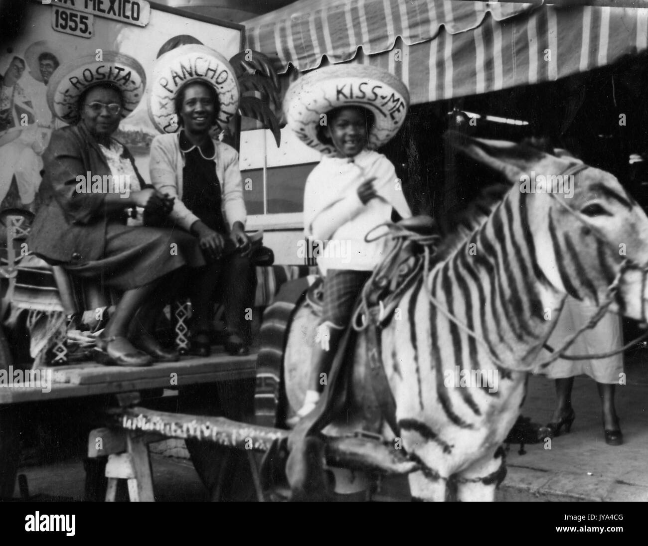 Afrikanische amerikanische Familie in Mexiko, zwei Frauen sitzen auf einem Wagen, junges Kind reiten einen Esel, lackiert ist, sich wie ein Zebra Look, alle tragen Sombreros, 1955. Stockfoto