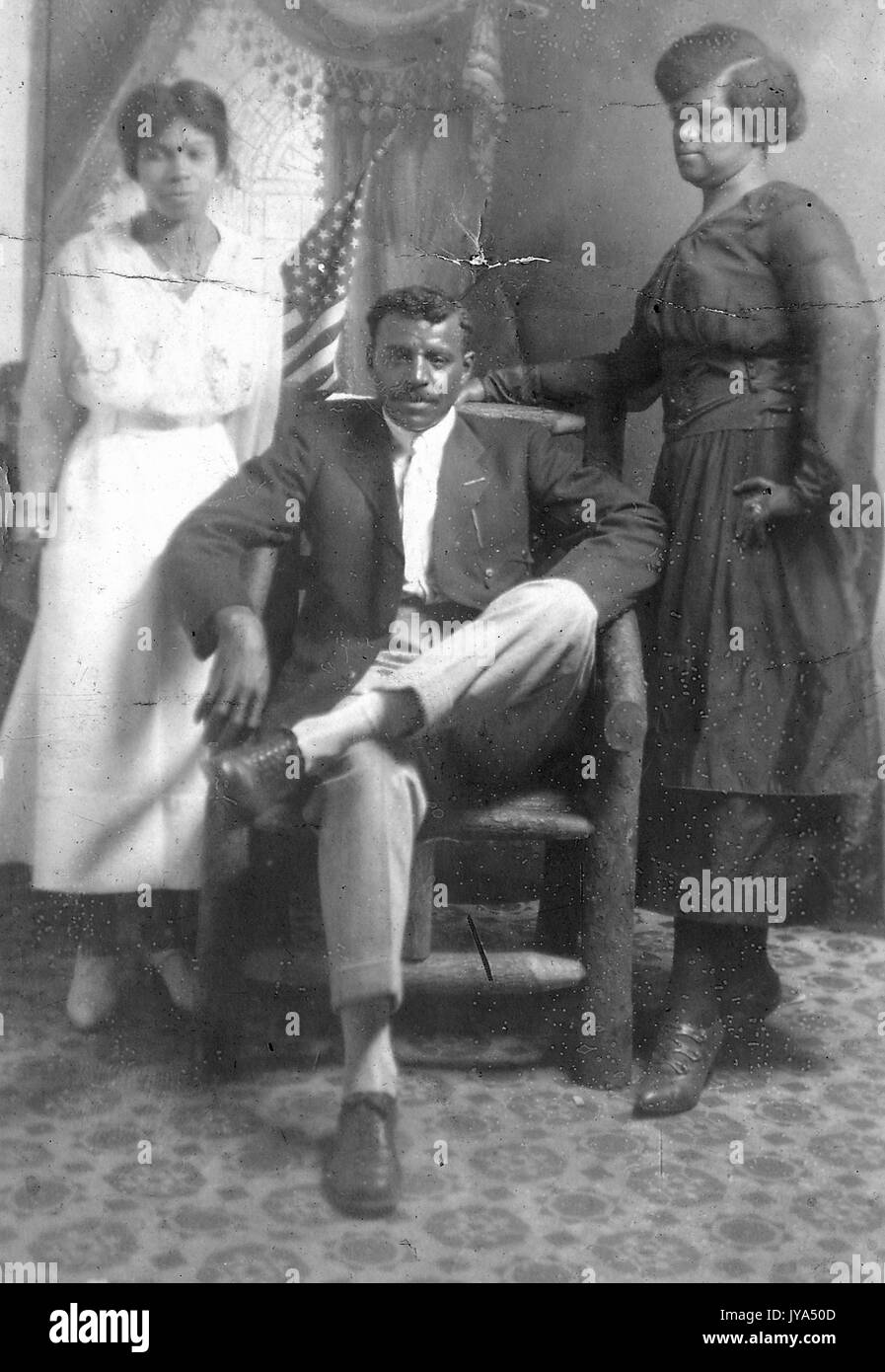 Porträt eines afrikanischen amerikanischen Familie, der Vater in einem Stuhl sitzend mit der Mutter steht ihre Hand ruht auf dem Stuhl, Tochter stehen auf der linken Seite der Eltern, oder gemalten Hintergrund verfügt über eine amerikanische Flagge, 1910. Stockfoto