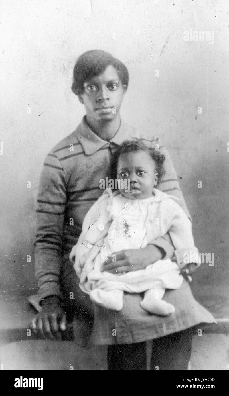 Afrikanische amerikanische Frau auf einer Bank sitzen und ihr Baby Holding, beide mit leeren Gesichtsausdruck und das Aussehen der Ausbauchende Augen, die Frau ihre Hand ruht auf der Bank, 1910. Stockfoto