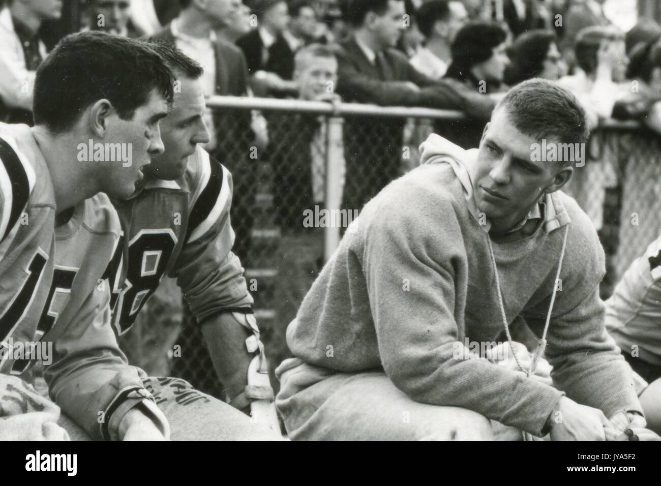 Ein junger Lacrosse-Spieler der Johns Hopkins University, der ein Sweatshirt auf seiner Uniform trägt, sitzt auf einer Bank und spricht mit zwei uniformierten Teamkollegen, einer Menge Zuschauer, die hinter einem Zaun im Hintergrund stehen, 1960. Stockfoto