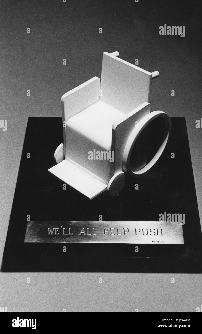Ein Miniatur-Rollstuhl auf einer Holztafel mit dem Titel "WIR WERDEN ALLE HELFEN ZU SCHIEBEN", MSE Symposium, 1970. Stockfoto