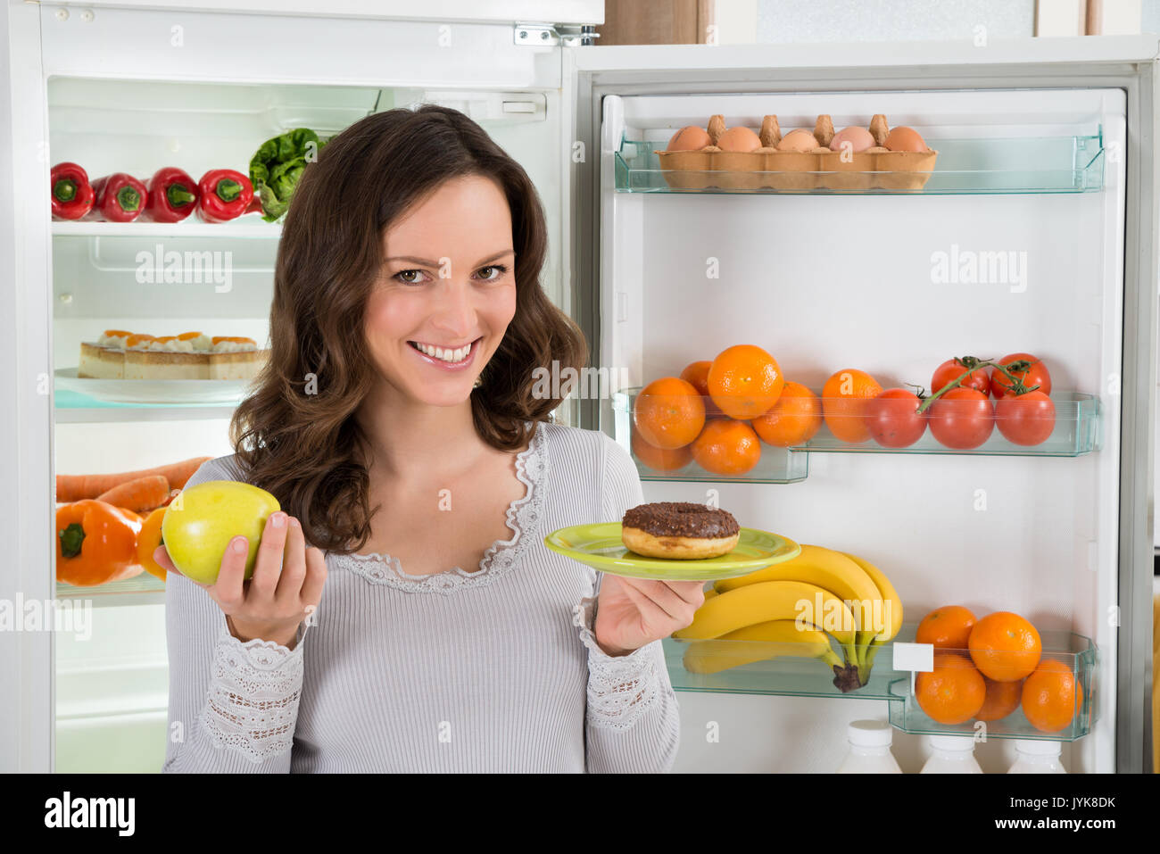 Junge Frau mit grünem Apfel und Krapfen Vor Kühlschrank Stockfoto