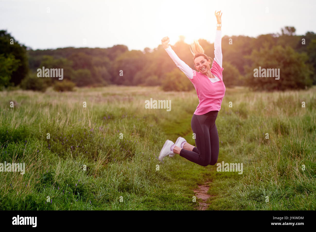 Passen glückliche junge Frau in Sportbekleidung springen in die Luft auf dem Weg durch Wiese Stockfoto