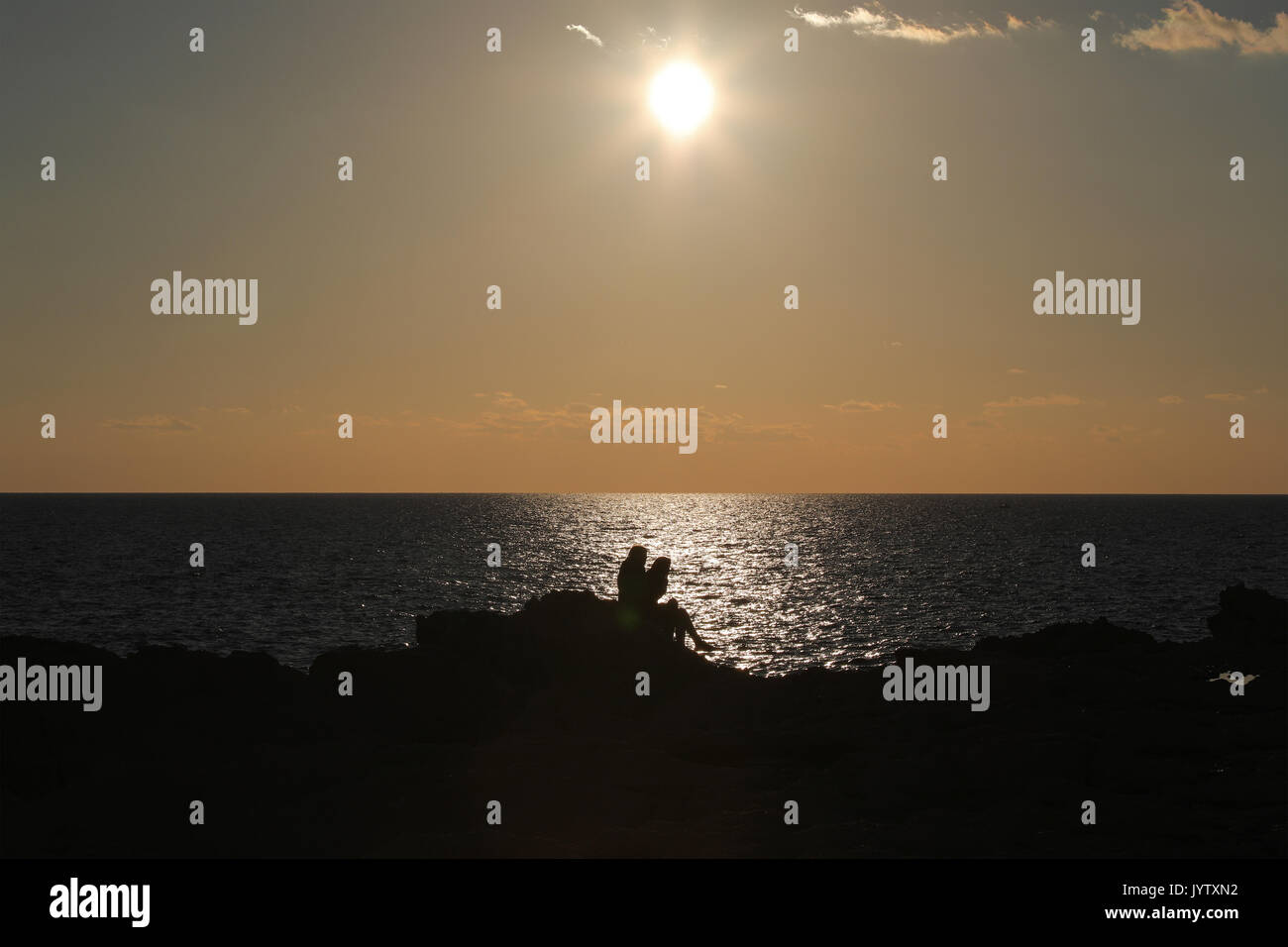 Silhouetten von zwei Personen sitzen auf Felsen am Meer bei Sonnenuntergang Stockfoto