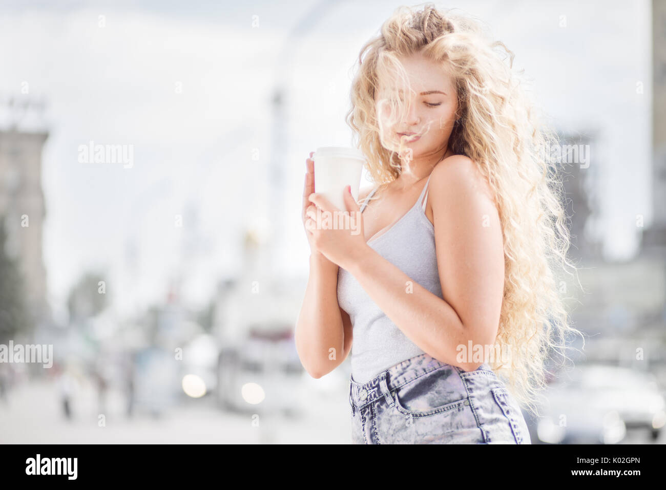 Glückliche junge Frau mit einer Tasse Kaffee zum Mitnehmen und lächelnd mit Flirt vor der Kamera gegen urban Stadtverkehr Hintergrund. Stockfoto