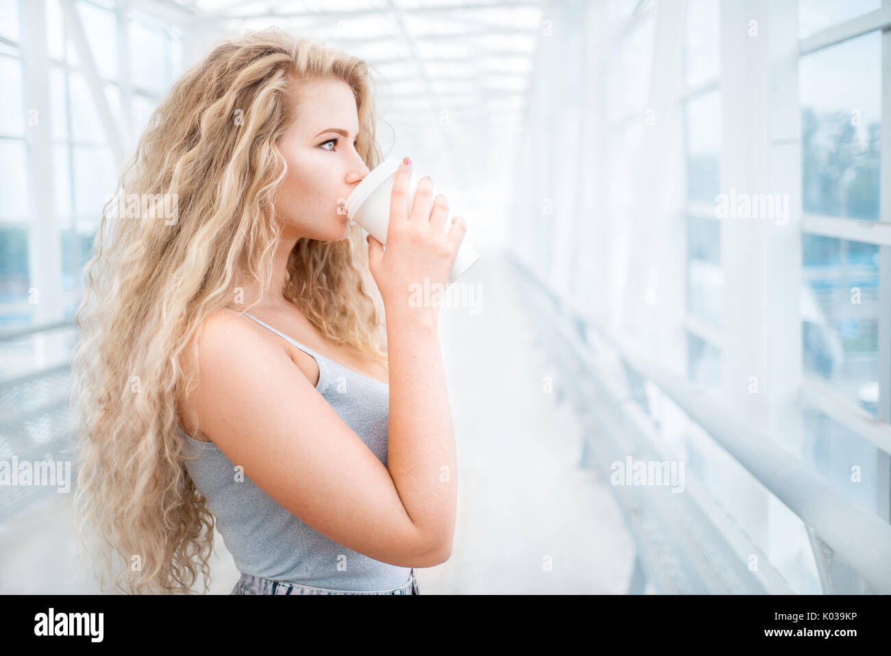 Schöne junge Frau mit langen lockigen Haaren, ein Take away Kaffeetasse halten und stehen auf der Brücke gegen städtischen Hintergrund. Stockfoto