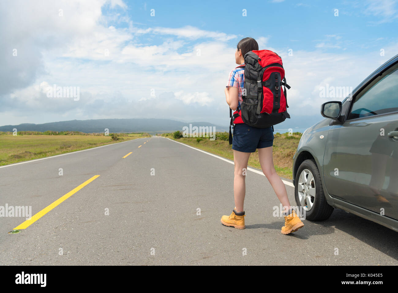 Elegante gemächliche weibliche Backpacker auf Asphalt beobachten entfernte Bergkulisse und geparkte Autos in der Straße am Sommer, Urlaub reisen. Stockfoto