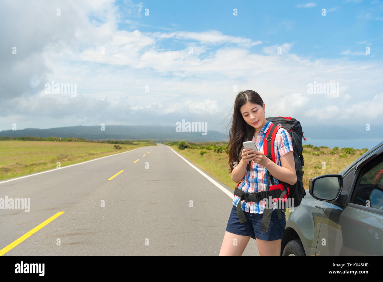 Lächelnd attraktiven weiblichen Touristen neben der Verwendung von Mobile Smartphone auf der Suche nach touristischen Informationen und Durchführung Rucksack stehendes Auto zu reisen Stockfoto