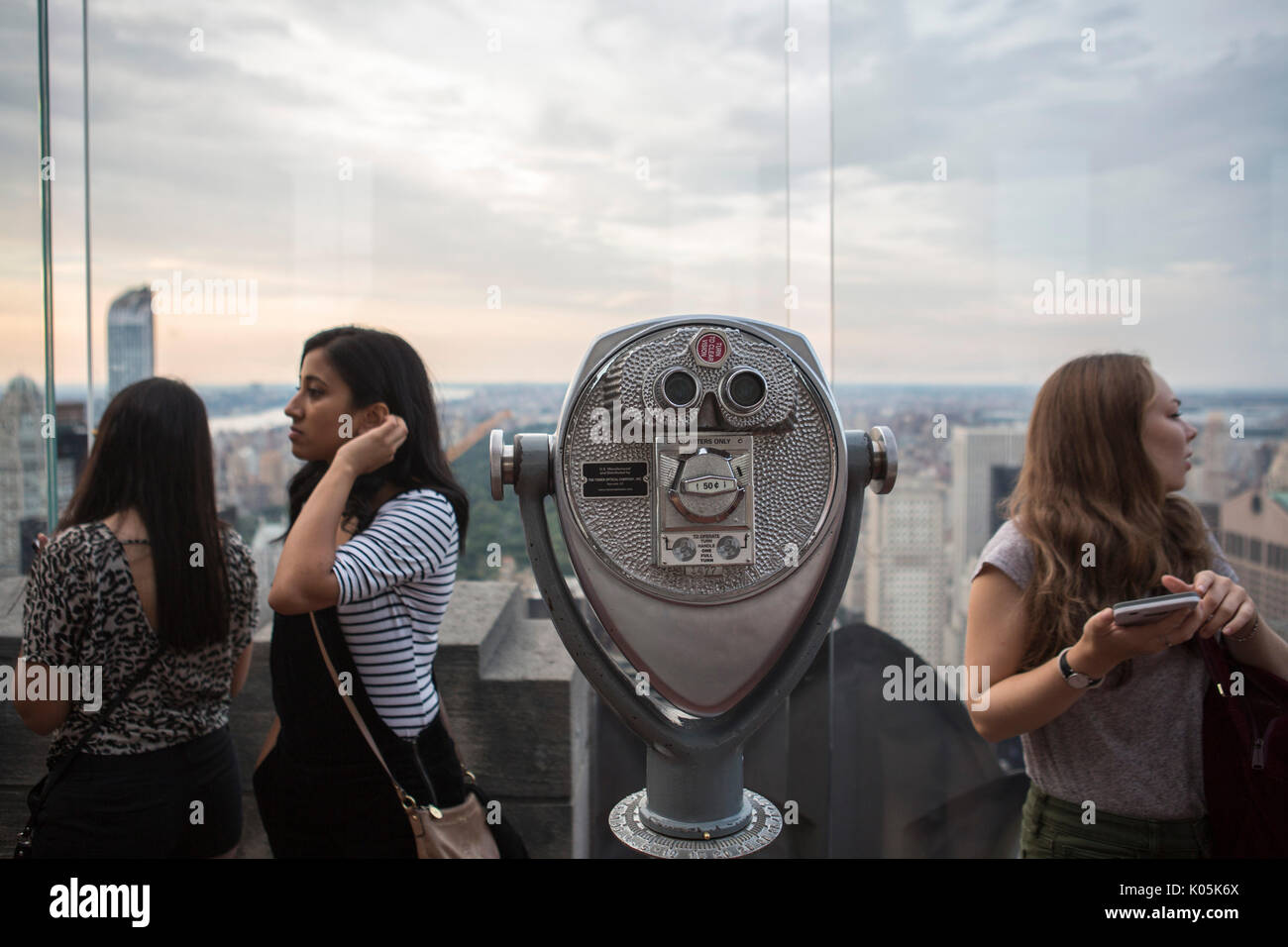 Touristen, die Foto's und selfies auf der Oberseite des Rockefeller Center mit Blick auf Downtown Manhattan mit dem Empire State Building, die Sonnenuntergänge über New York City und eine alte bezahlt Münzautomaten Teleskop im Vordergrund. Stockfoto