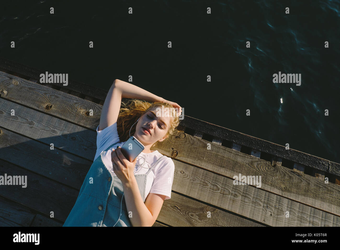 Junge schöne blonde Mädchen Teenager liegen auf Pier, dem Meer und Musik zu hören. Junge Frau mit Kopfhörer und Telefon outdoo Stockfoto