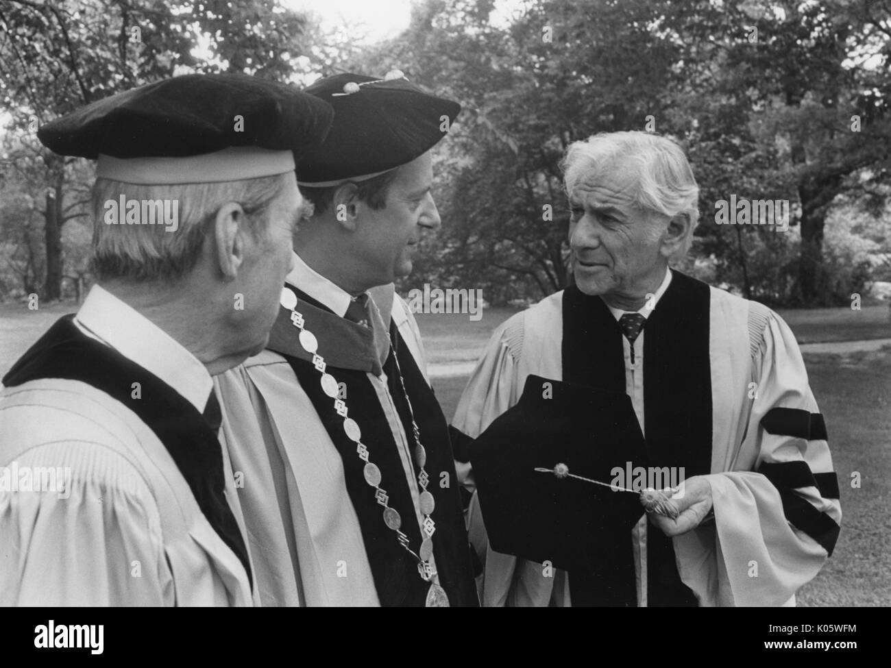 Brustbild von zwei Johns Hopkins Professoren und der Präsident, Steven Muller, mit amerikanischer Komponist und Pianist Leonard Bernstein auf seinem Recht, alle drei Männer in Kappen und Kleid geschmückt, stehend auf dem Gras außerhalb, Bernstein seinen Hut aus- und halten Sie es in der Hand, ernste Mimik, 1980. Stockfoto