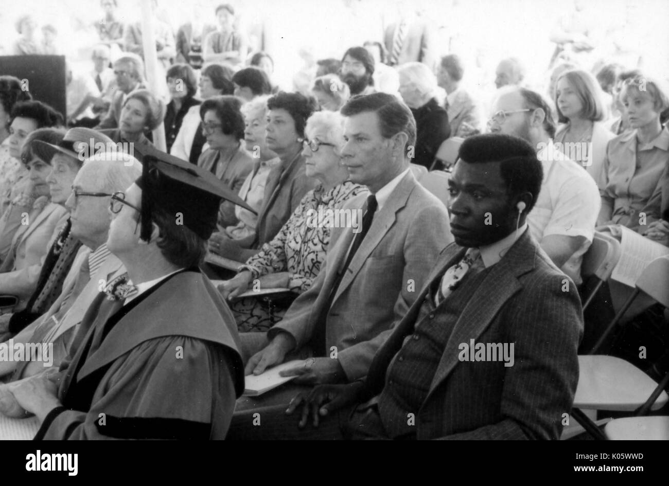 Portrait des Publikums an der Johns Hopkins Beginn sitzen, sitzen auf Plastikstühlen, eine afrikanische amerikanische Mann im Anzug im Vordergrund mit einer Hörmuschel in seinem linken Ohr, hinter einem Mitglied der Fakultät mit Kappe und Kleid sitzt, alle mit Blick auf die Anfang Zeremonie, mit ernsten Gesichtsausdruck, 1980. Stockfoto
