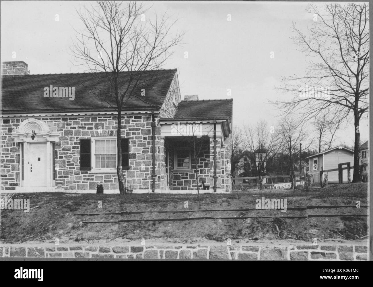 Landschaft geschossen von einem Teil ein einstöckiges Haus aus Stein, mit einer weißen Tür, dunklen Fensterläden, durch Bäume ohne Blätter umgeben, Roland Park/Boston, USA, 1910. Dieses Bild wird von einer Reihe dokumentieren den Bau und den Verkauf von Wohnungen in der Roland Park/Guilford Nachbarschaft von Baltimore, einer Straßenbahn Vorort und eines der ersten geplanten Gemeinschaften in den Vereinigten Staaten. Stockfoto