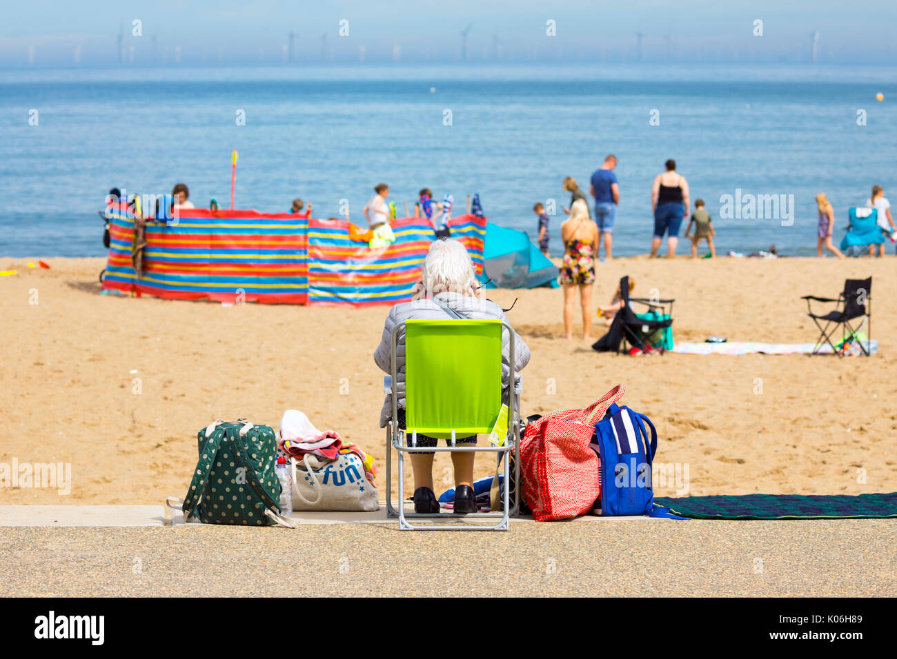 Ältere Frau saß alleine auf einer Liege mit ihren Taschen mit Blick auf einen überfüllten Strand im beliebten Badeort Colwyn Bay im Norden von Wales auf einem Sommertag während der Sommerferien, Wales, Großbritannien Stockfoto