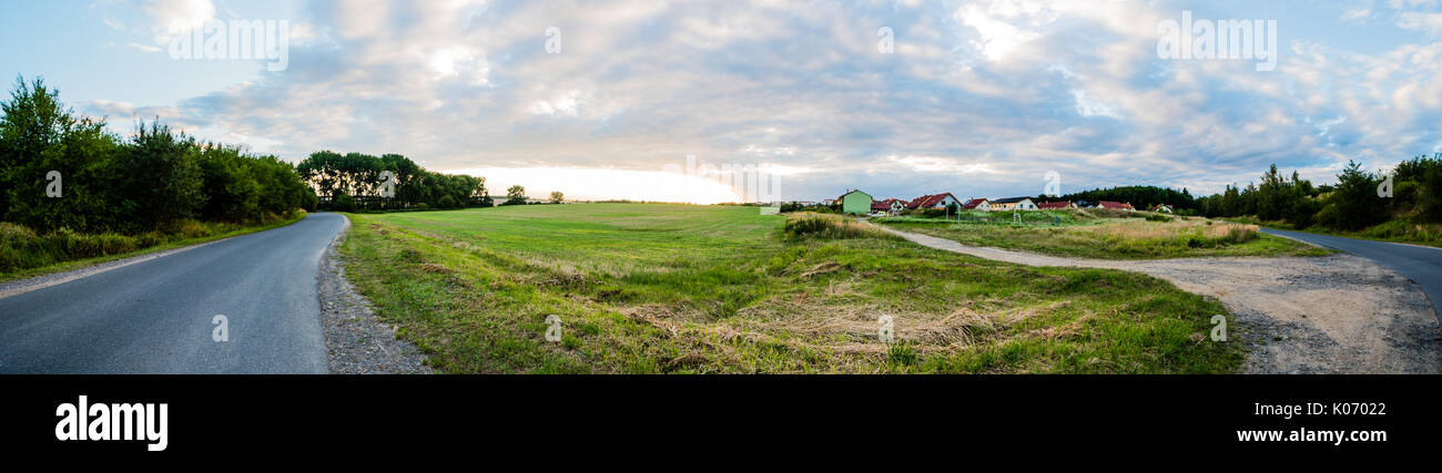 Landschaft Landschaft mit einigen Häusern in der kleinen Stadt mit Straße um. Grüne Wiese mit Bäumen und Gras, Sonnenuntergang am Horizont. Panoramablick. Nic Stockfoto