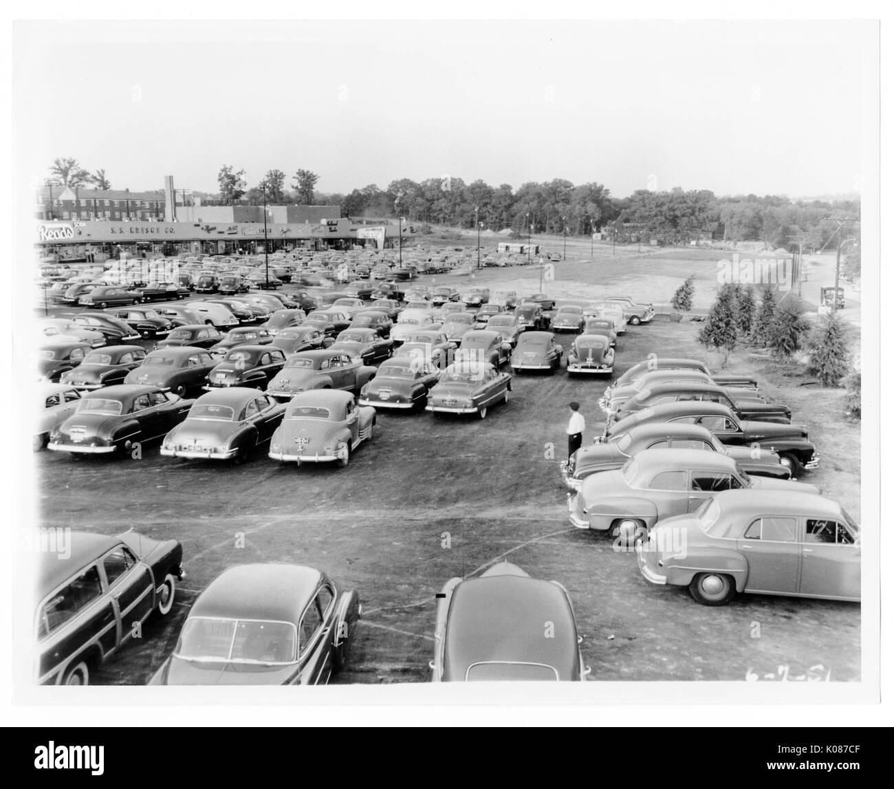 Parkplatz von Northwood Einkaufszentrum mit Hunderten von geparkten Autos und Bäumen und Laternen im Hintergrund, Stores wie Liest auf der Rückseite, Mann, der Parkplatz neben Autos, Baltimore, Maryland, 1950. Stockfoto