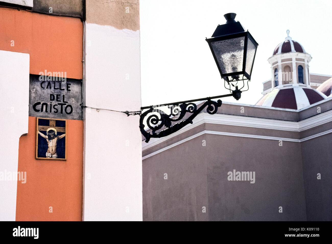 Eine religiöse Kunstwerke von keramischen Fliesen zeigt Jesus Christus am Kreuz und markiert die Calle del Cristo (Straße, in der sich das Kruzifix), führt zu der Kathedrale von San Juan Bautista (rechts) in der historischen Altstadt San Juan in Puerto Rico (PR), ein gemeindefreies Gebiet der Vereinigten Staaten in der Karibik. Ein vintage Schmiedeeisen Straße Licht der Straße Name aufleuchtet und illustrierte Zeichen in der Nacht. Stockfoto