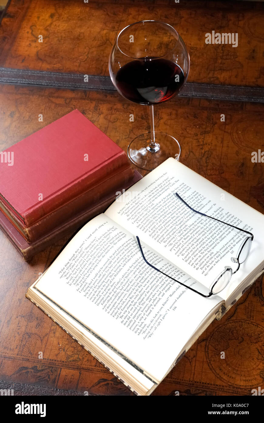 Ein offenes Buch auf einem Leder Couchtisch sitzt neben einem Stapel Bücher und einem Glas Rotwein. Lesebrille auf dem geöffneten Seiten. Stockfoto