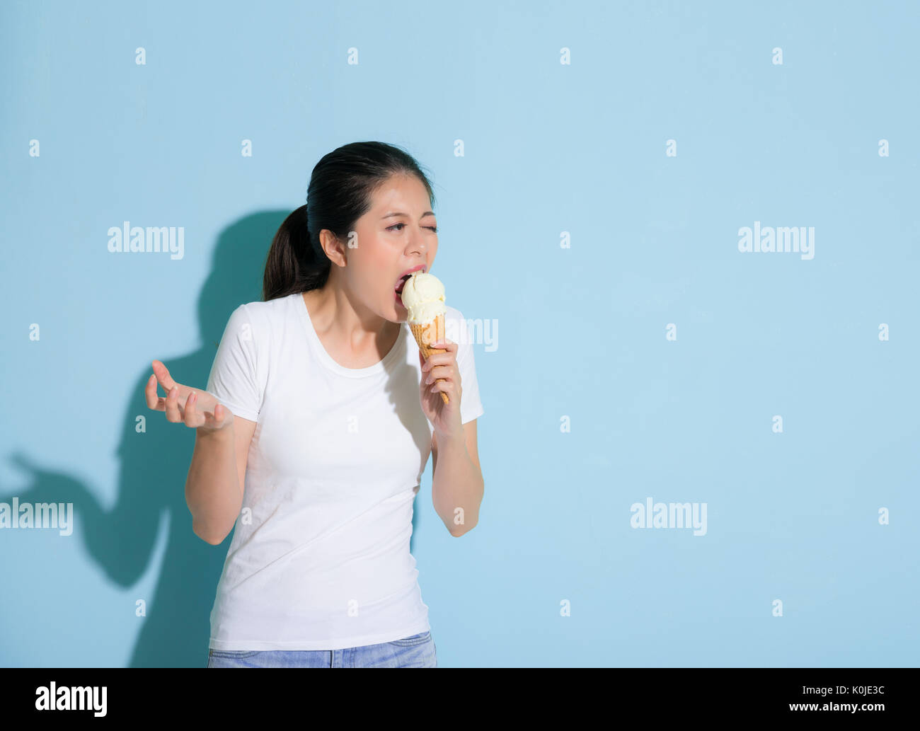 Süße attraktives Mädchen öffnen großen Mund essen Vanilleeis und Gesten Greifer Gefühl empfindliche Zähne schmerzhaft Leistung in blaue Wand backgr Stockfoto