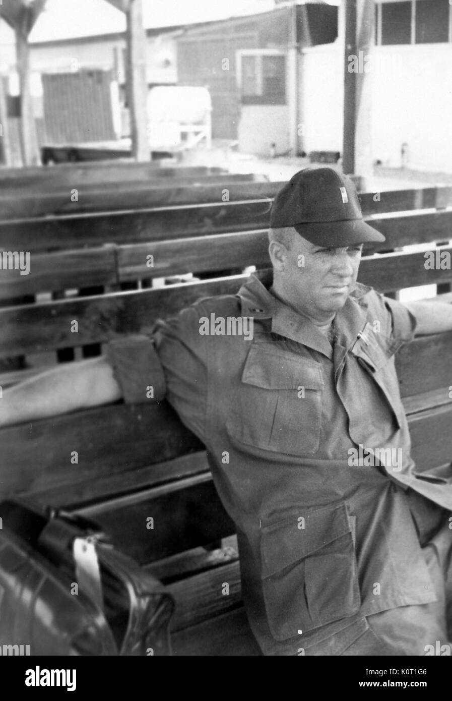 Soldat, trug einen Baseball Cap, auf einer Bank sitzen, die Arme auf den Seiten über die Spitze, mit Blick in die Ferne, Zeilen von Bänken, die hinter ihm, Vietnam, 1964. Stockfoto