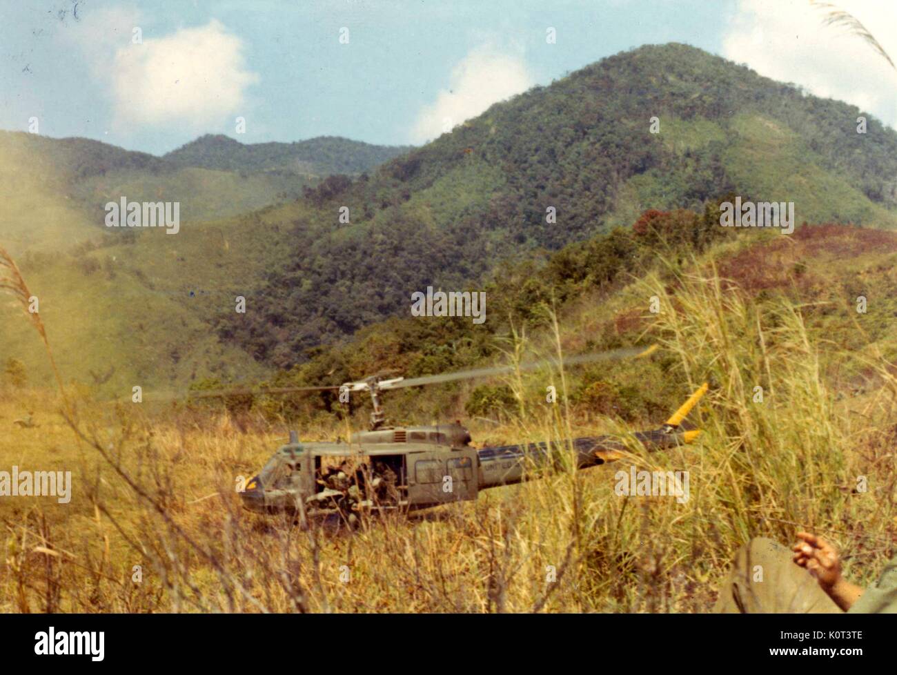 Huey Hubschrauber Landung in einem Feld in Vietnam während des Vietnam Krieges, Marines Aussteigen aus dem Hubschrauber, der Bein und Hand eines amerikanischen Soldaten im Vordergrund das Rauchen einer Zigarette, 1965 sichtbar. Stockfoto
