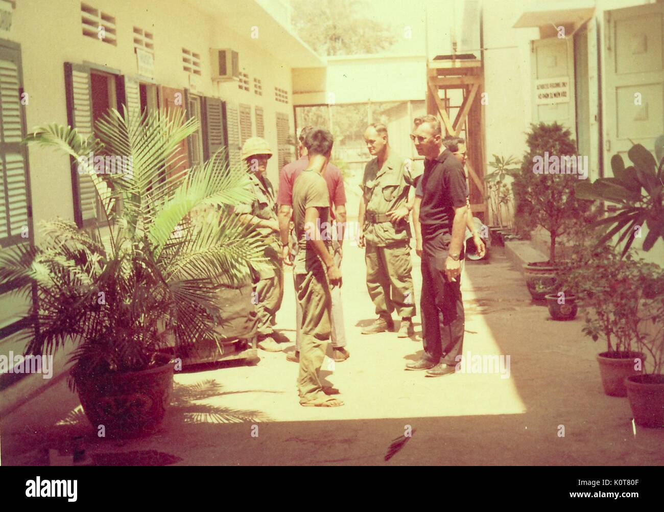 Ein Foto von einer Gruppe von Menschen sprechen miteinander, zwei der Männer sind gekleidet in United States Army Combat Uniform, die anderen Männer lässig gekleidet sind, sind Sie im Innenhof zwischen zwei Gebäuden versammelt und von Topfpflanzen, Vietnam, 1968 umgeben. Stockfoto