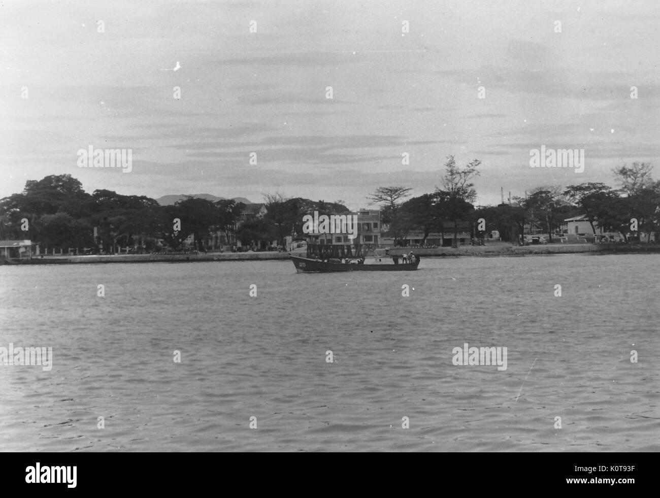 Ein langes Boot fährt entlang eines Flusses, wo Häuser in der Nähe der Küste, Vietnam, 1966 erbaut wurden. Stockfoto