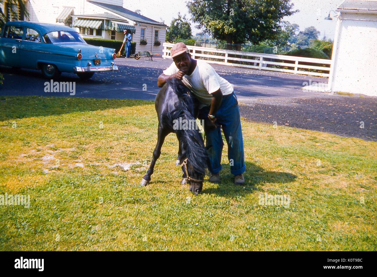 Portrait einer afrikanischen amerikanischen Mann mit einer jungen Schwarzen gezügelten Pferd in einem grasbewachsenen Innenhof Posing, Juni, 1959. Ein blaues Auto in der Einfahrt hinter ihm geparkt. Im Hintergrund, einem anderen afrikanischen amerikanischen Mann fegt Blätter außerhalb einer schalbrett Haus. Stockfoto